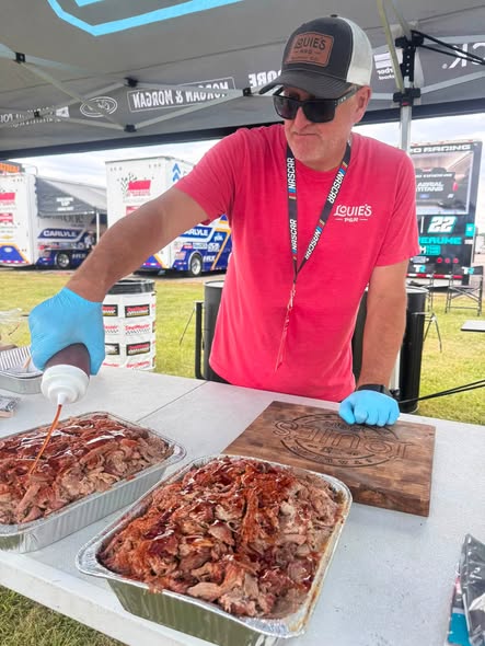 A Louie's pitmaster in gloves saucing trays of pulled pork at a race-weekend cook